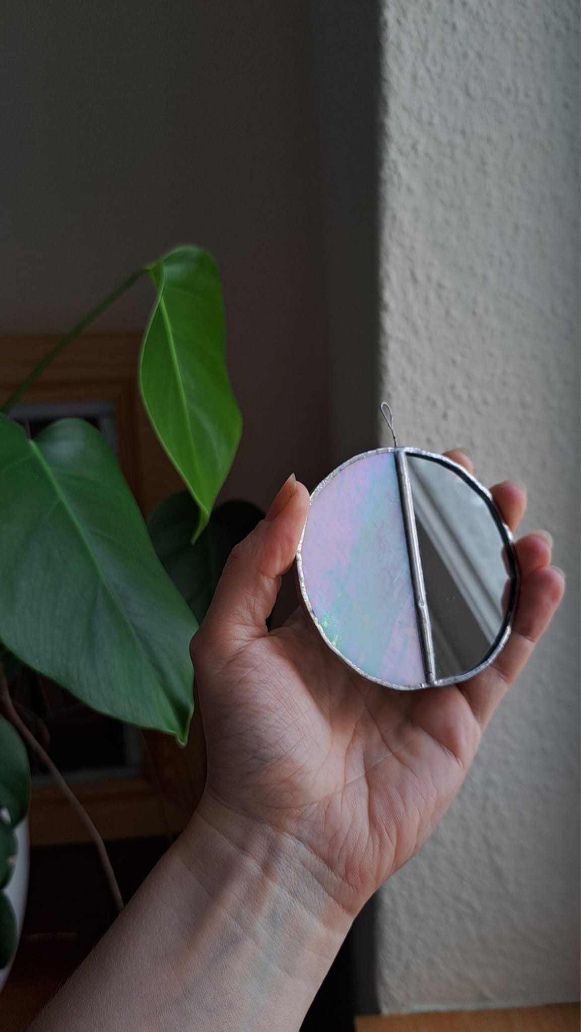 A hand holding a small, circular, iridescent mirror with a silver metal frame, set against a background of a textured wall and a green houseplant.