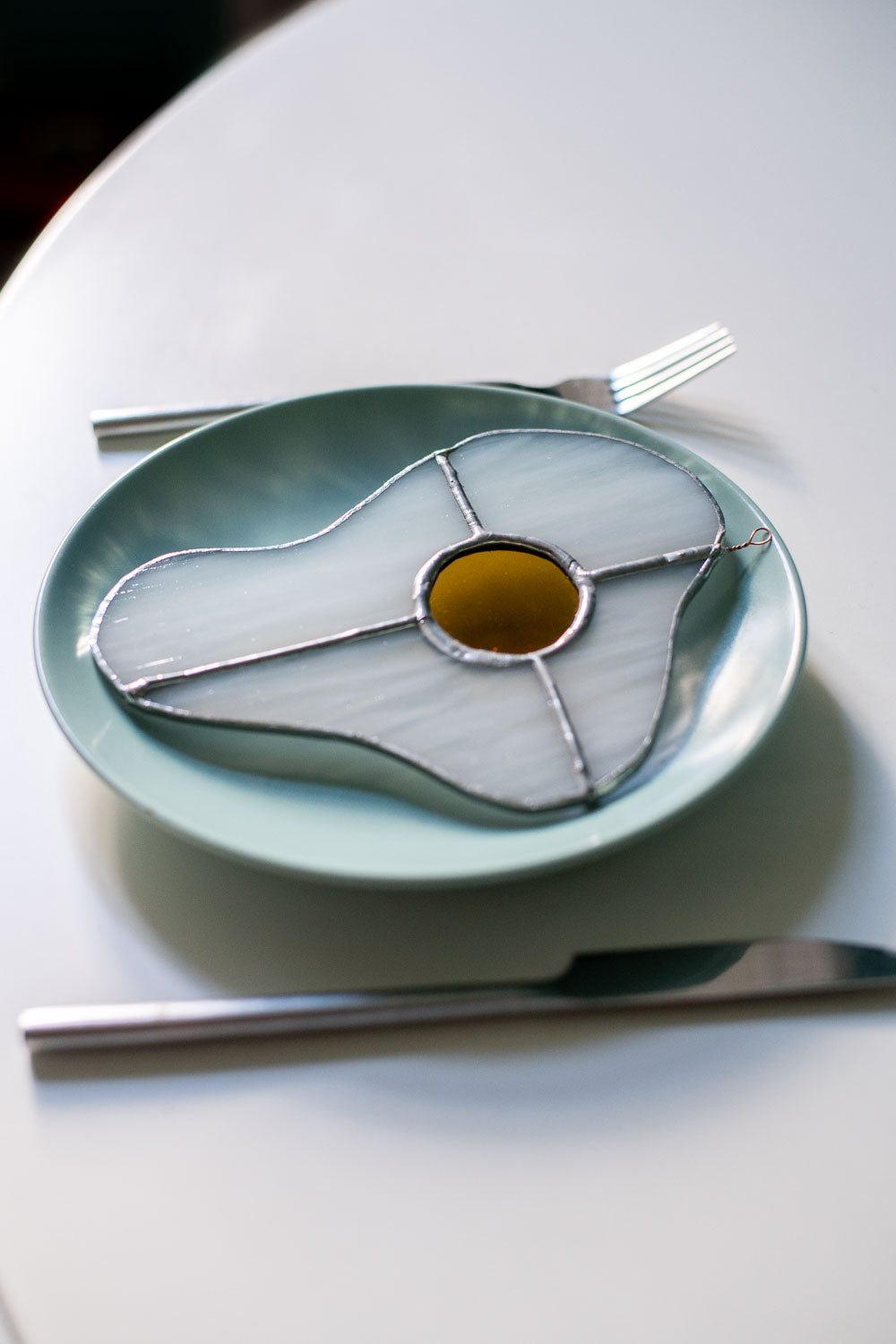 Teal plate with a decorative egg-shaped stained glass artwork, fork, knife, and glass on a white surface.