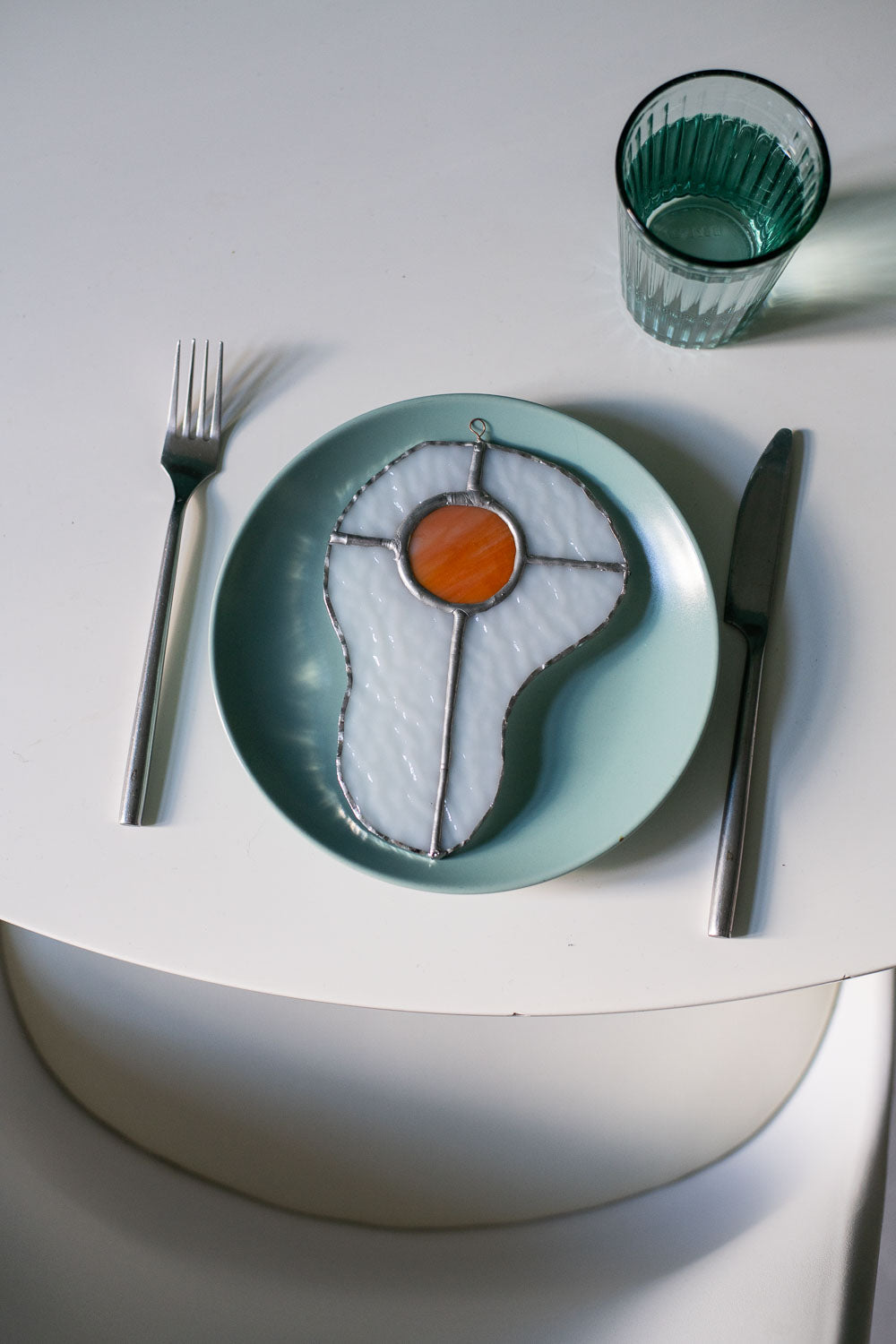 Teal plate with a decorative egg-shaped stained glass artwork, fork, knife, and glass on a white surface.
