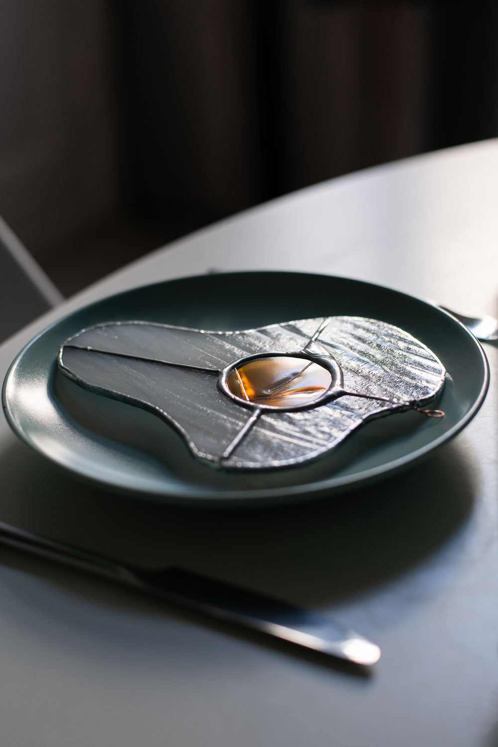 A stained glass art work in the shape of a sunny side up egg on a blue ceramic plate on a whitetable with a glass and cutlery.