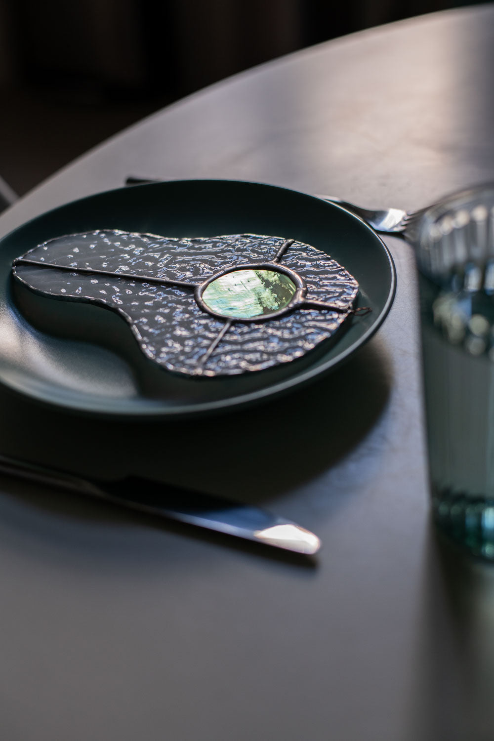 Teal plate with a decorative egg-shaped stained glass artwork, fork, knife, and glass on a white surface.
