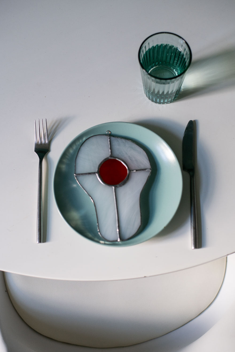 Teal plate with a decorative egg-shaped stained glass artwork, fork, knife, and glass on a white surface.