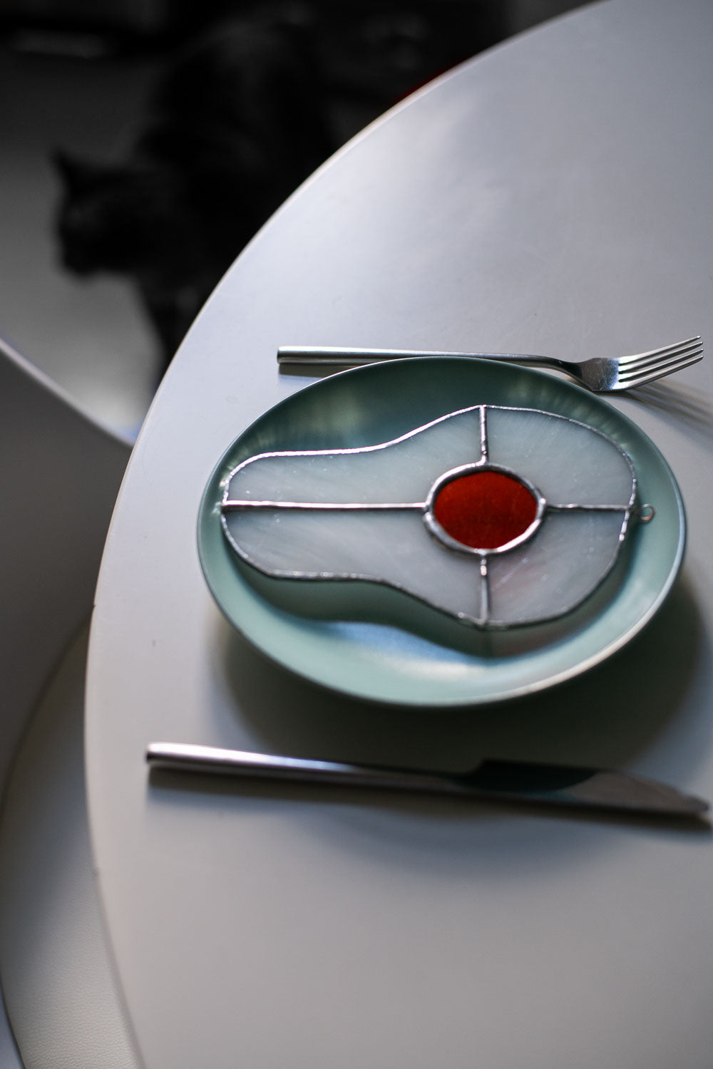 Teal plate with a decorative egg-shaped stained glass artwork, fork, knife, and glass on a white surface.