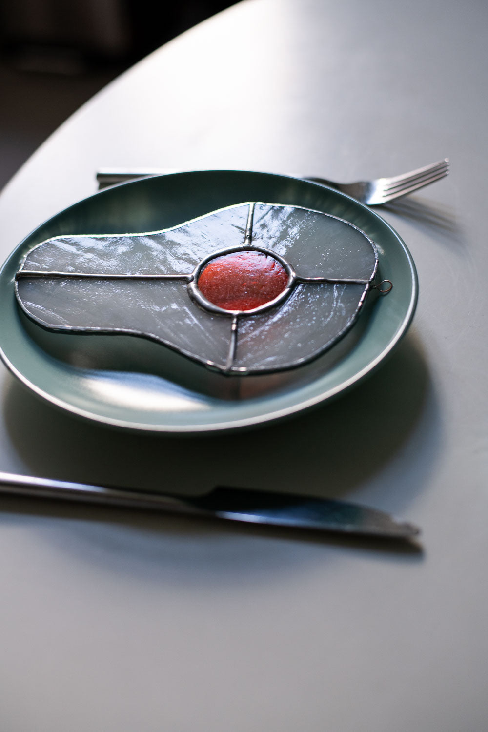 Teal plate with a decorative egg-shaped stained glass artwork, fork, knife, and glass on a white surface.
