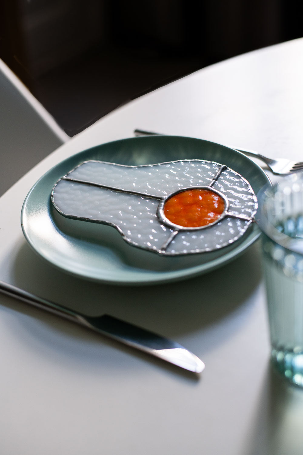 Teal plate with a decorative egg-shaped stained glass artwork, fork, knife, and glass on a white surface.