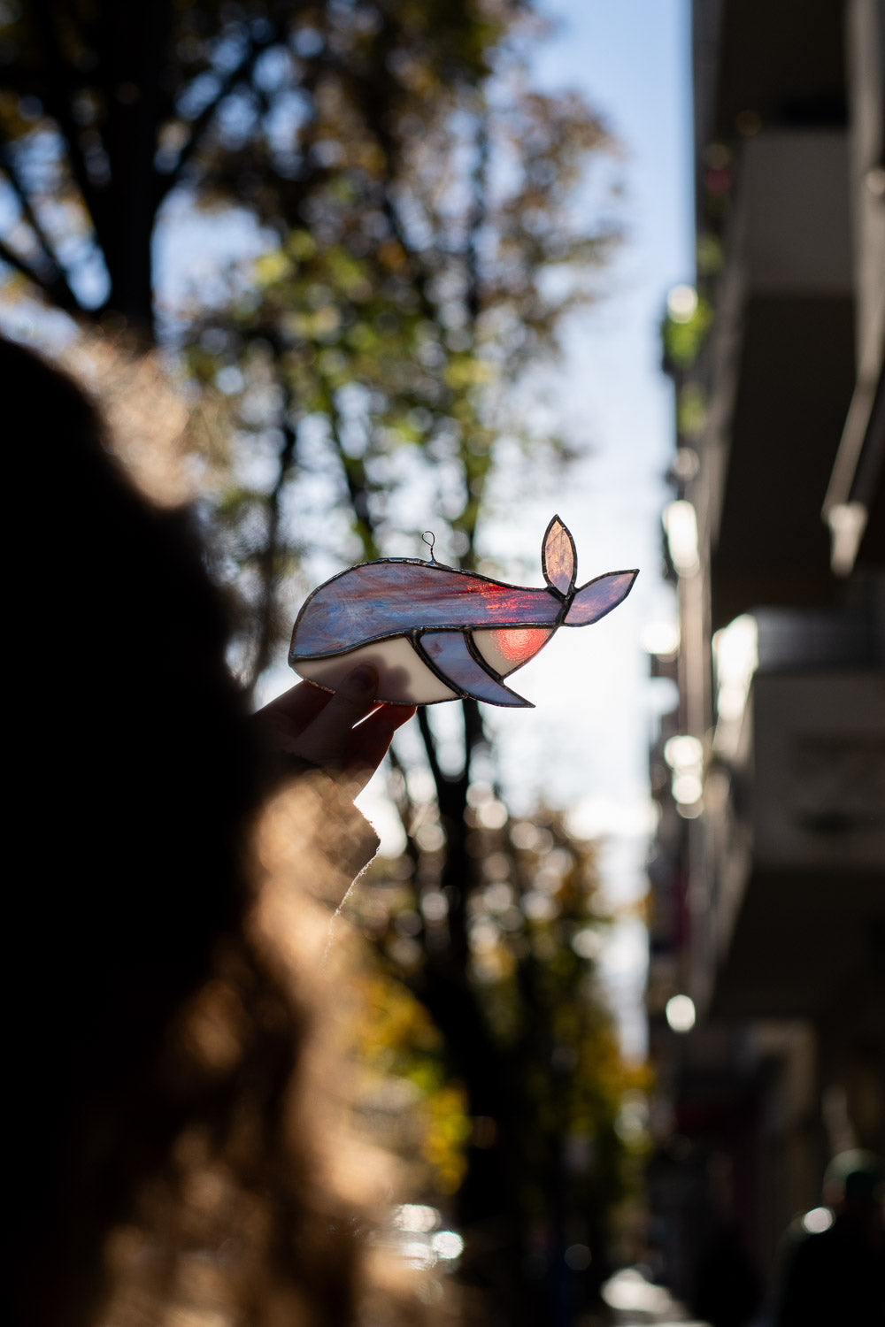 Decorative stained glass piece in the shape of a whale held by a person with trees and buildings in the background