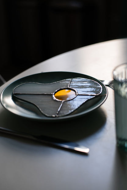 Teal plate with a decorative egg-shaped stained glass artwork, fork, knife, and glass on a white surface.